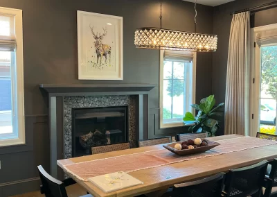 A modern dining room featuring a wooden table, black chairs, and a pendant light is elevated by the touch of skilled interior painters. The fireplace boasts an artwork of a stag above the mantel, creating a cozy yet elegant atmosphere.