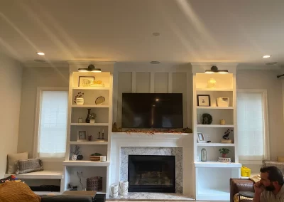 A cozy living room featuring a fireplace, TV, and artfully decorated shelves. The expertly painted walls, courtesy of Alpha Omega Finishers, complement the inviting seating area and warm lighting that beautifully illuminate the space. interior painting