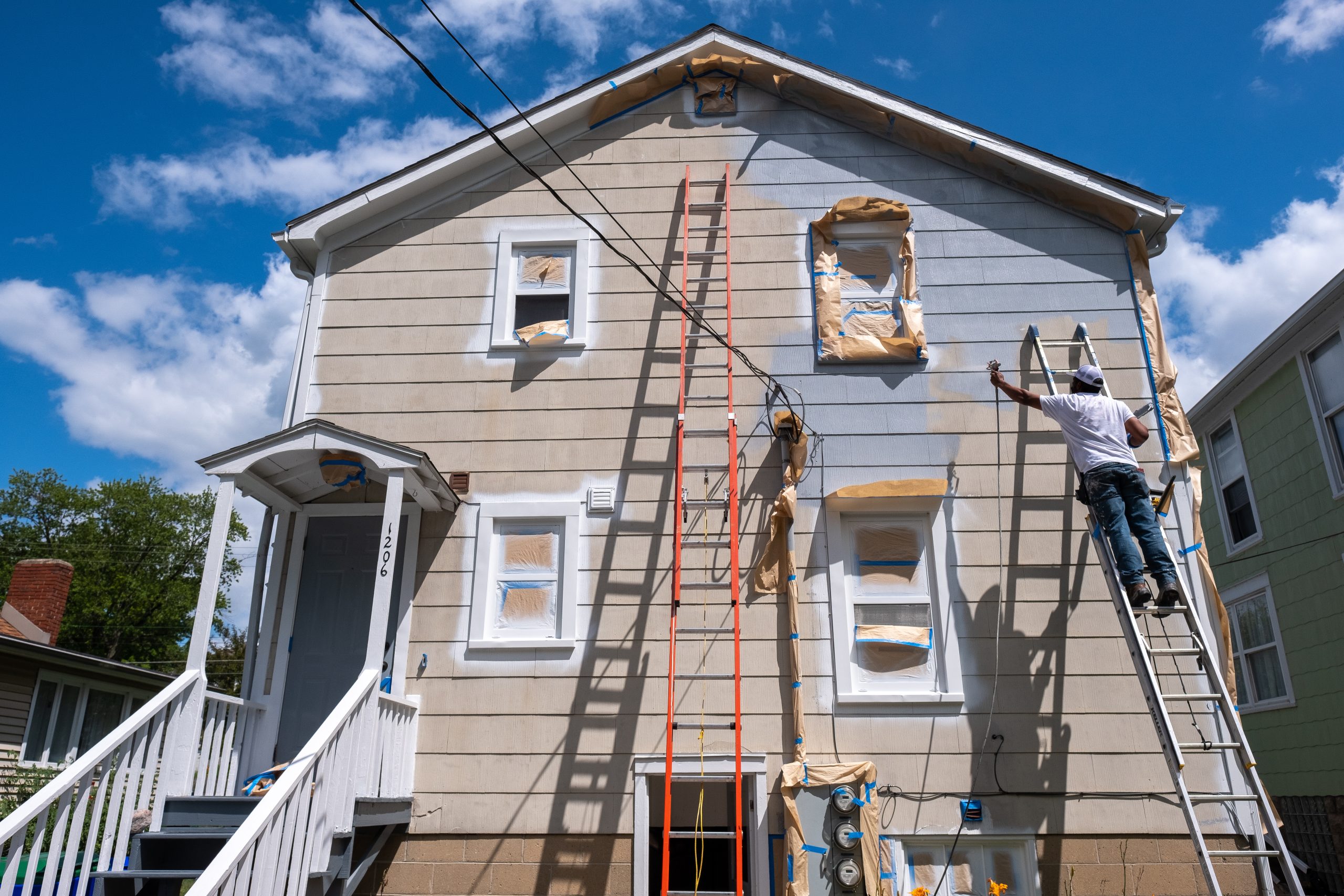 Painter on a ladder spraying paint on the exterior of a house A person on a ladder performs professional exterior painting, coating the two-story house beneath a blue sky with clouds.