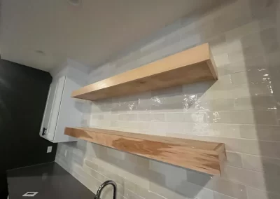 Two wooden floating shelves grace the white tiled kitchen wall above a black countertop and sink, showcasing Alpha Omega Finishers' exquisite attention to detail.