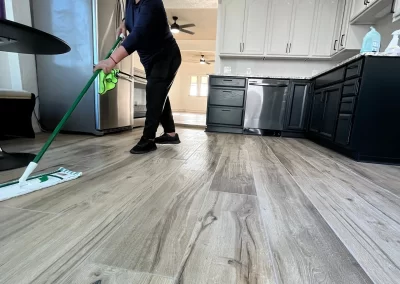 A person mops the kitchen floor of this beautifully maintained space, featuring white cabinets and stainless steel appliances. The wood flooring gleams, showcasing the attention to detail that Alpha Omega Finishers brings to every project, whether it's a spotless clean or expert house painting.