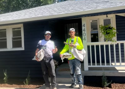 Two people standing in front of a dark blue house, smiling with painting tools in hand; their joy evident as they prepare for a day of exterior painting. The lush greenery provides the perfect backdrop for the craftsmanship of Alpha Omega Finishers.