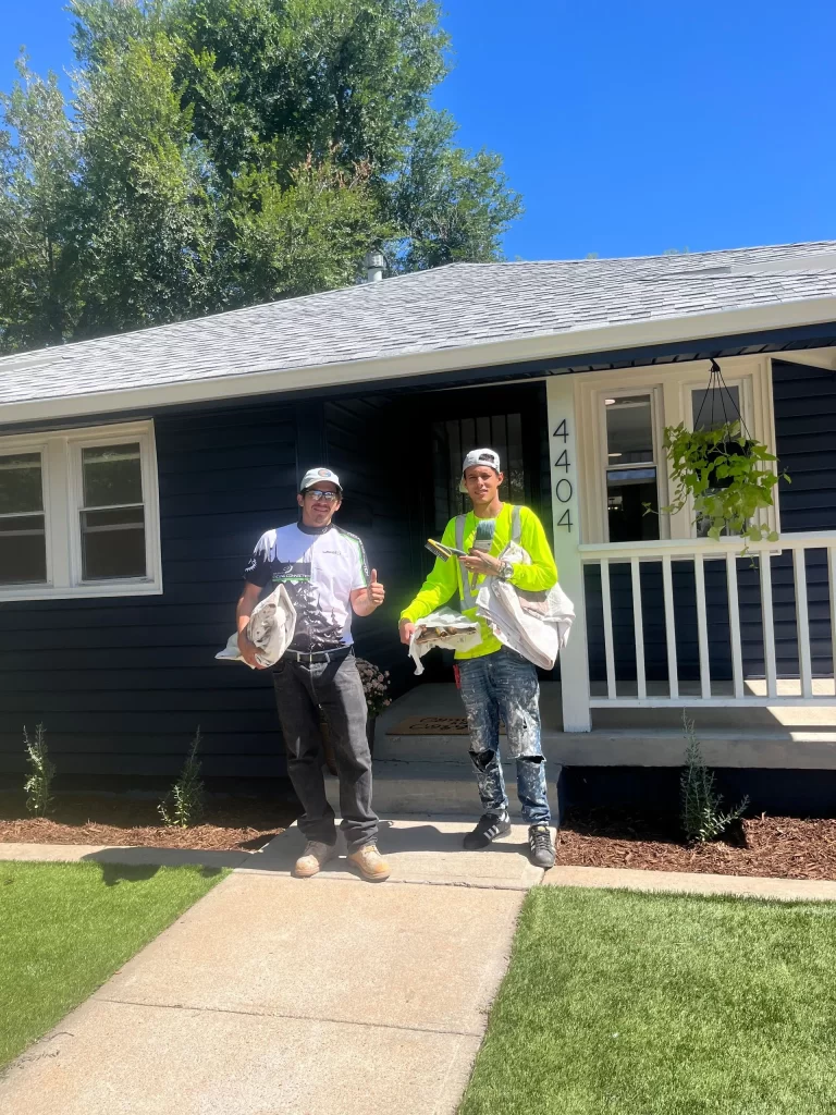 Two people standing in front of a dark blue house, smiling with painting tools in hand; their joy evident as they prepare for a day of exterior painting. The lush greenery provides the perfect backdrop for the craftsmanship of Alpha Omega Finishers.