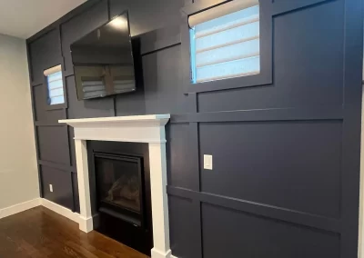 Discover a cozy living room featuring a dark accent wall crafted by Alpha Omega Finishers, complete with a TV, inviting fireplace, two bright windows, and elegant hardwood floors.