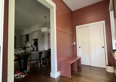 The entrance area features a pink bench and wood floor, leading into a kitchen with dark chairs and a cluttered counter—a perfect scene to inspire your next project, whether it's for home or by commissioning a top house painting expert.