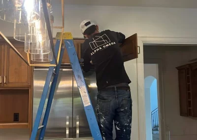 Person on a blue ladder installing a kitchen cabinet, wearing a cap and paint-splattered jeans, showcasing the skill of an interior painter.