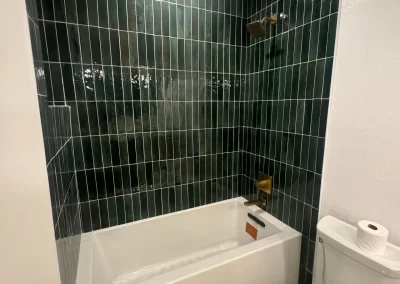 Bathroom with green tile walls, white tub, and modern toilet. Light gray patterned floor designed by interior painters and a stylish ceiling light complete the serene space.