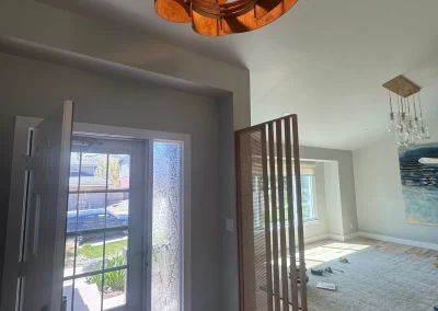 Interior entryway with wooden floor, circular chandelier, glass door, and open living area featuring a freshly painted wall by expert interior painters, accented by a large rug and window.