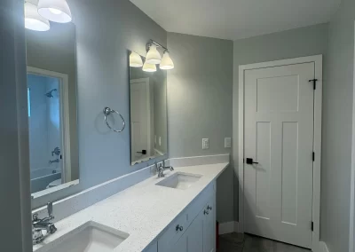 Modern bathroom with a long double-sink vanity, large mirror, stylish light fixtures, and a pristine white door. The light gray walls and tiled floor are expertly crafted, showcasing the touch of skilled interior painters.
