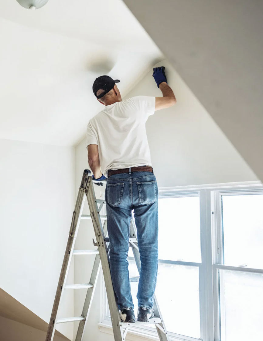 A person on a ladder wearing gloves, expertly  interior painting a white wall in a room with a large window. With the precision of Alpha Omega Finishers, this house painting project transforms the space with meticulous care and attention to detail.