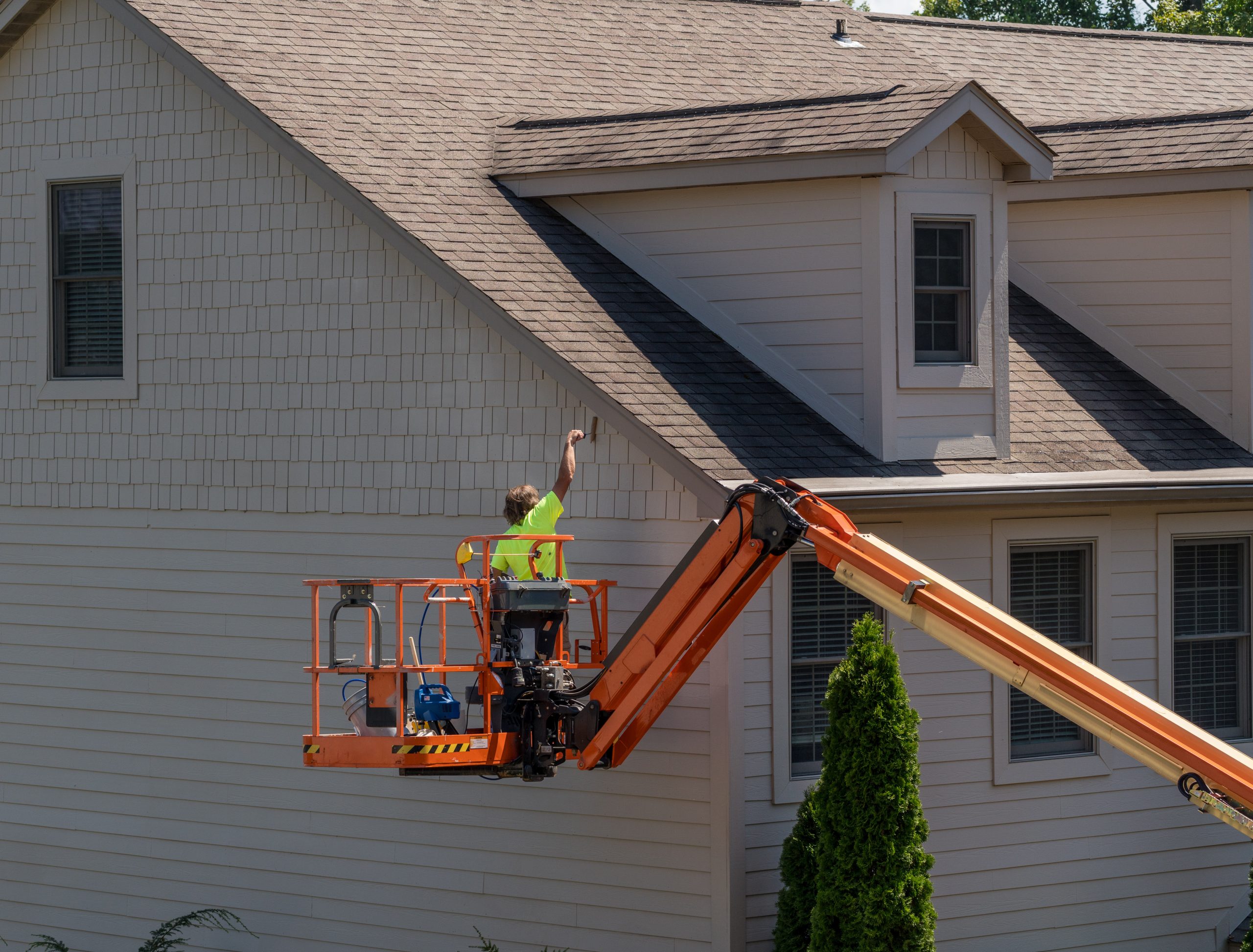 House painter in articulating boom lift paints side of home Person on an orange lift paints siding of a beige house with multiple windows and a steep roof.