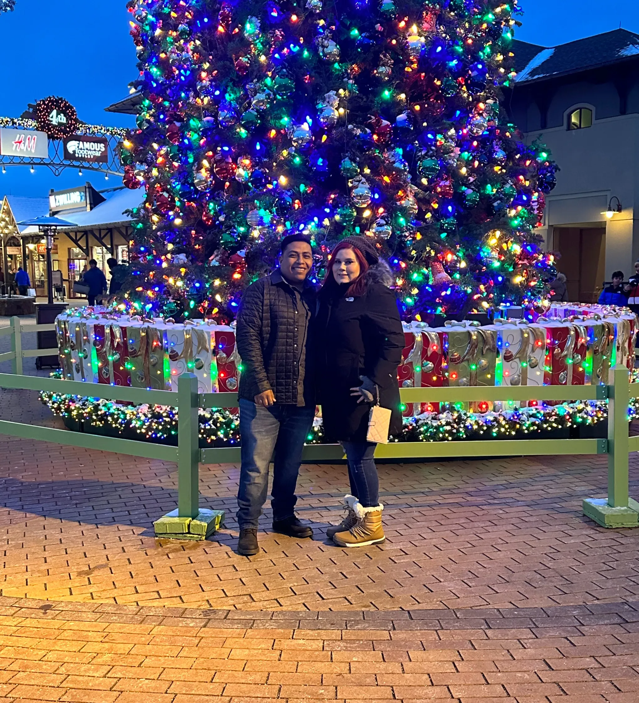 A couple stands smiling in front of a large, brightly lit Christmas tree outdoors in the evening.