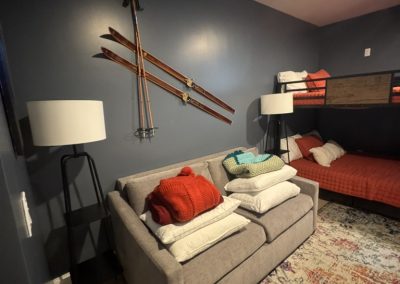 Cozy room with a gray sofa, pillows, vintage skis on the wall, and a red bed with a bunk above.