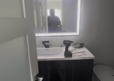 Modern bathroom with a lit mirror, white sink, tools on the counter, and a person reflected in the mirror.