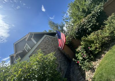 An American flag hangs on a brick house surrounded by green plants and a sunny blue sky.