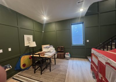A playroom with green walls, a table with chairs, toys, a rug, and a window with blinds.