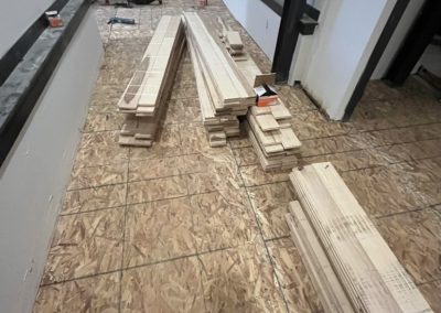 Person in red shirt installing wood flooring; wooden planks stacked on unfinished floor in a room.