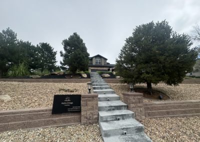 Gray house at the top of a concrete staircase, surrounded by trees and rocks, with a black sign in front.
