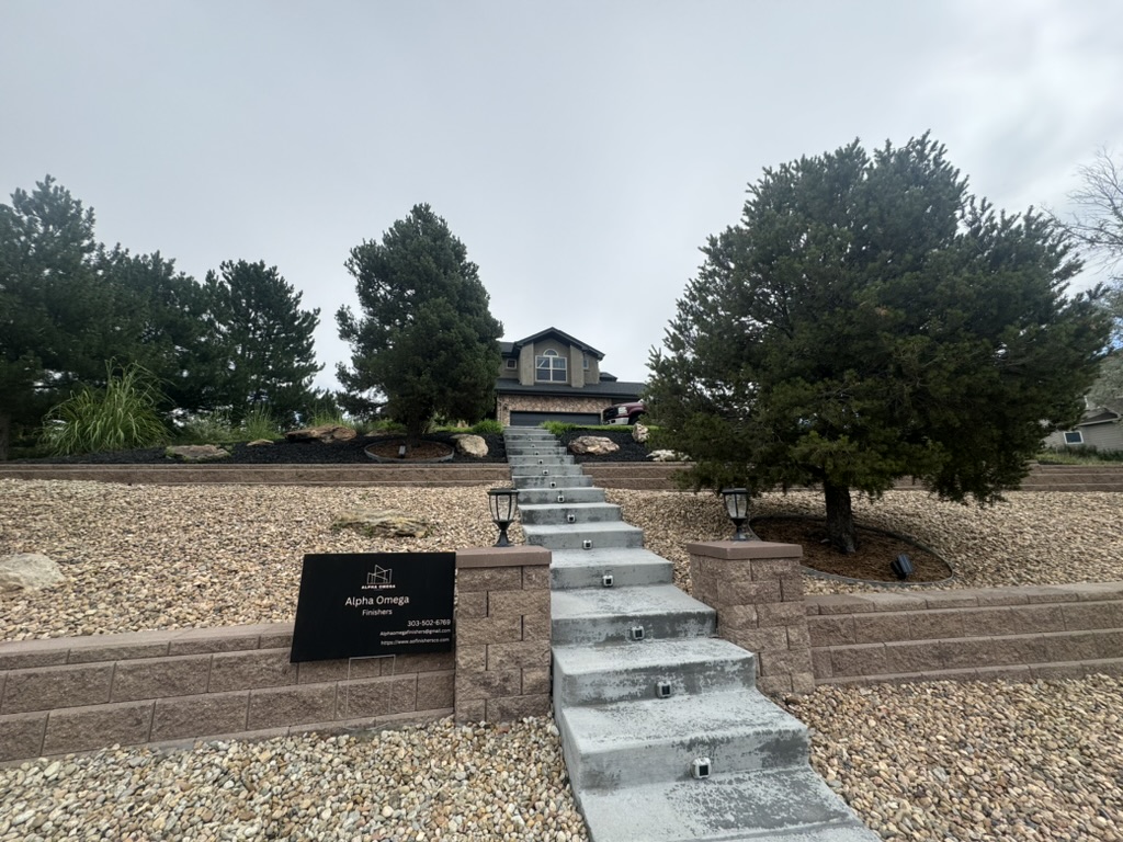 Gray house at the top of a concrete staircase, surrounded by trees and rocks, with a black sign in front.
