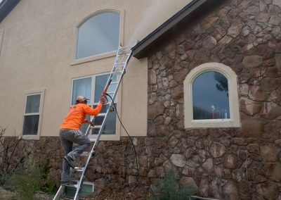 Person in an orange jacket power washing the upper wall of a house while standing on a ladder.