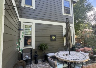 Sunny patio with a round table, chairs, umbrella, plants, and outdoor decor beside a two-story house.