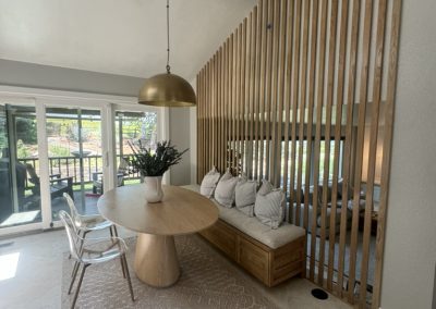 Modern dining nook with a round wooden table, built-in bench, slatted divider, and large windows.