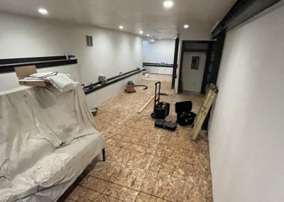A long, unfinished room with plywood flooring, tools, and covered furniture along white walls.