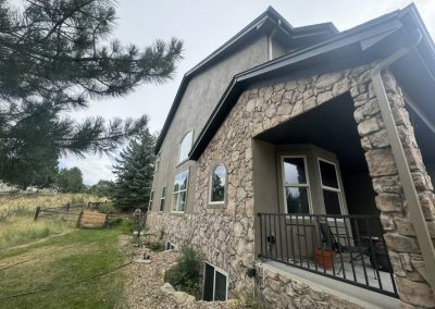 Two-story stone and stucco house with a small porch, surrounded by grass, trees, and a wooden fence.