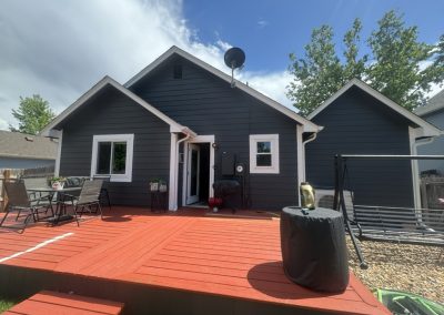 Backyard with red wooden deck, black house siding, patio furniture, grill, and covered swing set on a sunny day.