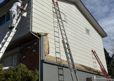 A person on a ladder paints the exterior wall of a two-story house on a sunny day.