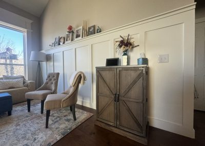 Living room with two tan chairs, a cabinet, and photos on a shelf above white wainscoting.
