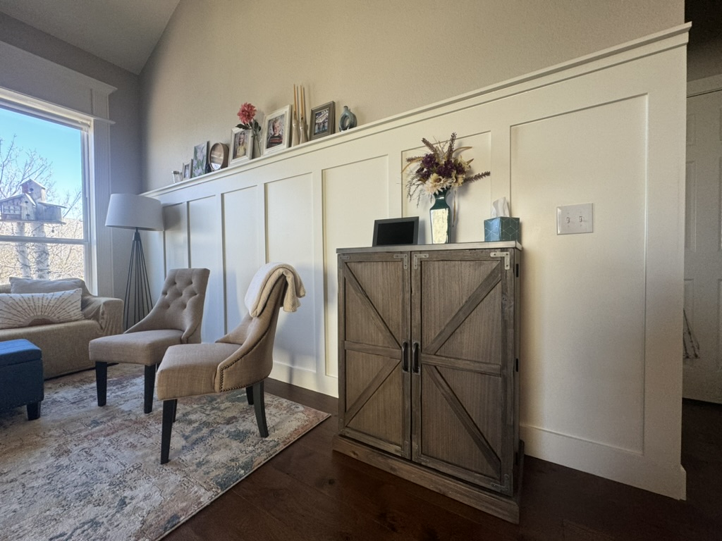 Living room with two tan chairs, a cabinet, and photos on a shelf above white wainscoting.