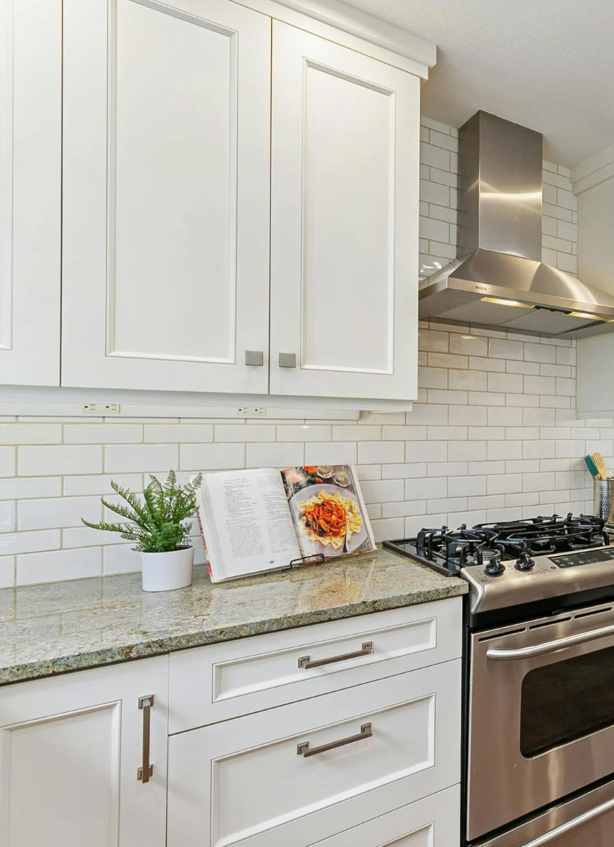 Modern kitchen with white cabinets, a gas stove, backsplash tiles, a plant, and an open cookbook on the counter.