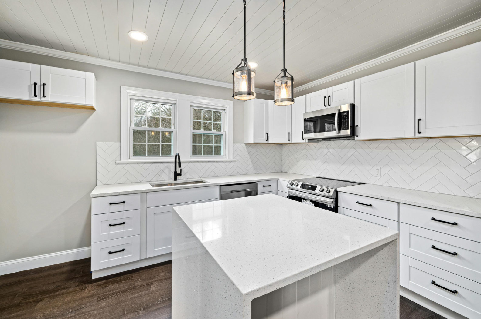 Modern white kitchen with island, stainless steel appliances, pendant lights, and herringbone tile backsplash.