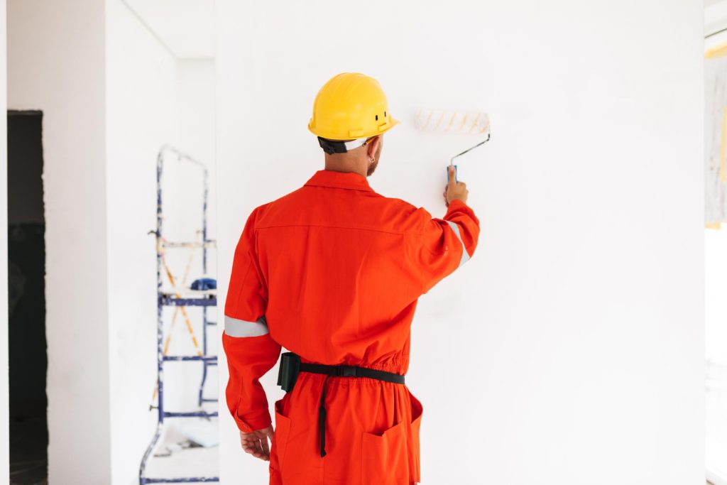 A worker in an orange uniform and yellow helmet paints a white wall with a paint roller.