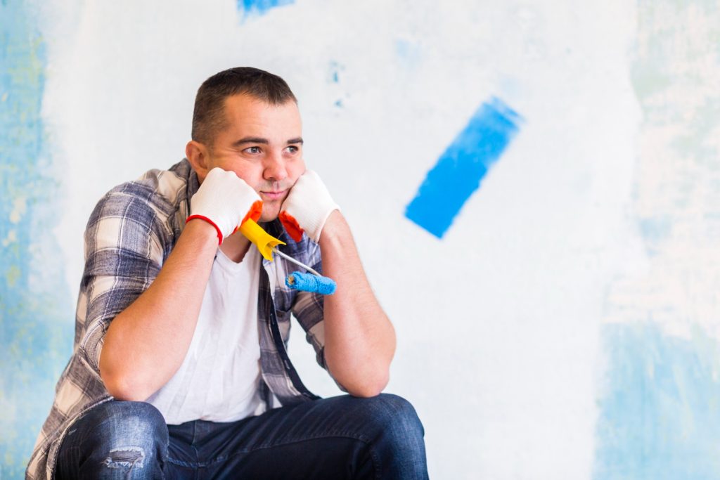 Man in gloves holding a paint roller sits and looks thoughtful, with blue paint patches on the wall behind him.