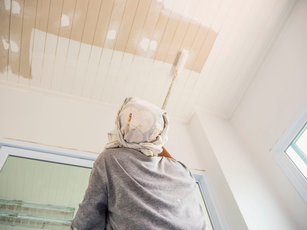 Person with a headscarf painting a white ceiling with a roller in a bright room.