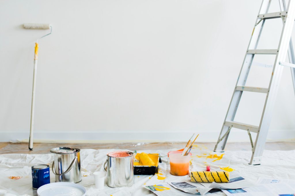 Paint cans, brushes, and a ladder set up for painting against a blank white wall with a roller and drop cloth.