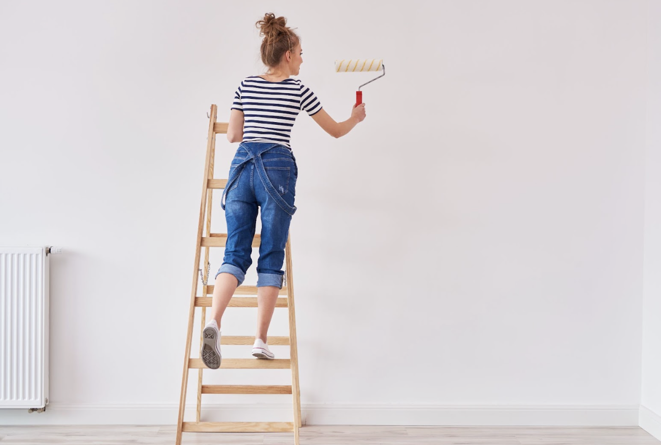 Professional House Painters vs DIY Denver A woman stands on a ladder painting a white wall with a roller in a bright room.
