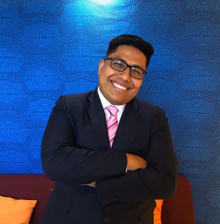 Smiling man in a suit with arms crossed stands in front of a blue American Express logo wall.